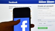 A person holds an iPhone displaying the Facebook app logo in front of a computer screen showing the facebook login page on August 3, 2016 in London, England.