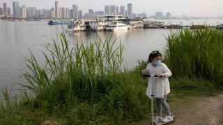 In this April 16, 2020, photo, a child wearing a mask against coronavirus rides her push bike along the banks of the Yangtze River in Wuhan in central China's Hubei province.
