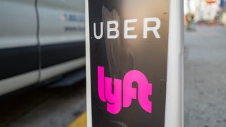 Close-up of vertical sign with logos for ridesharing companies Uber and Lyft, with wheels of a car in the background, indicating a location where rideshare pickups are available in downtown Los Angeles, California, October 24, 2018.