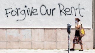 In this May 1, 2020, file photo, a woman wearing a mask walks past a wall bearing graffiti asking for rent forgiveness amid the COVID-19 pandemic in Los Angeles, California.
