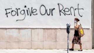 In this May 1, 2020, file photo, a woman wearing a mask walks past a wall bearing graffiti asking for rent forgiveness amid the COVID-19 pandemic in Los Angeles, California.