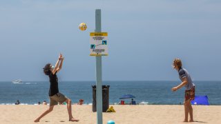 Two people wearing face masks play volleyball on the beach.