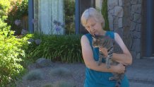 A woman holds a grey cat in her arms while standing in front of her home.