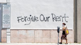 A woman wearing a mask walks past a wall bearing a graffiti asking for rent forgiveness on La Brea Ave.