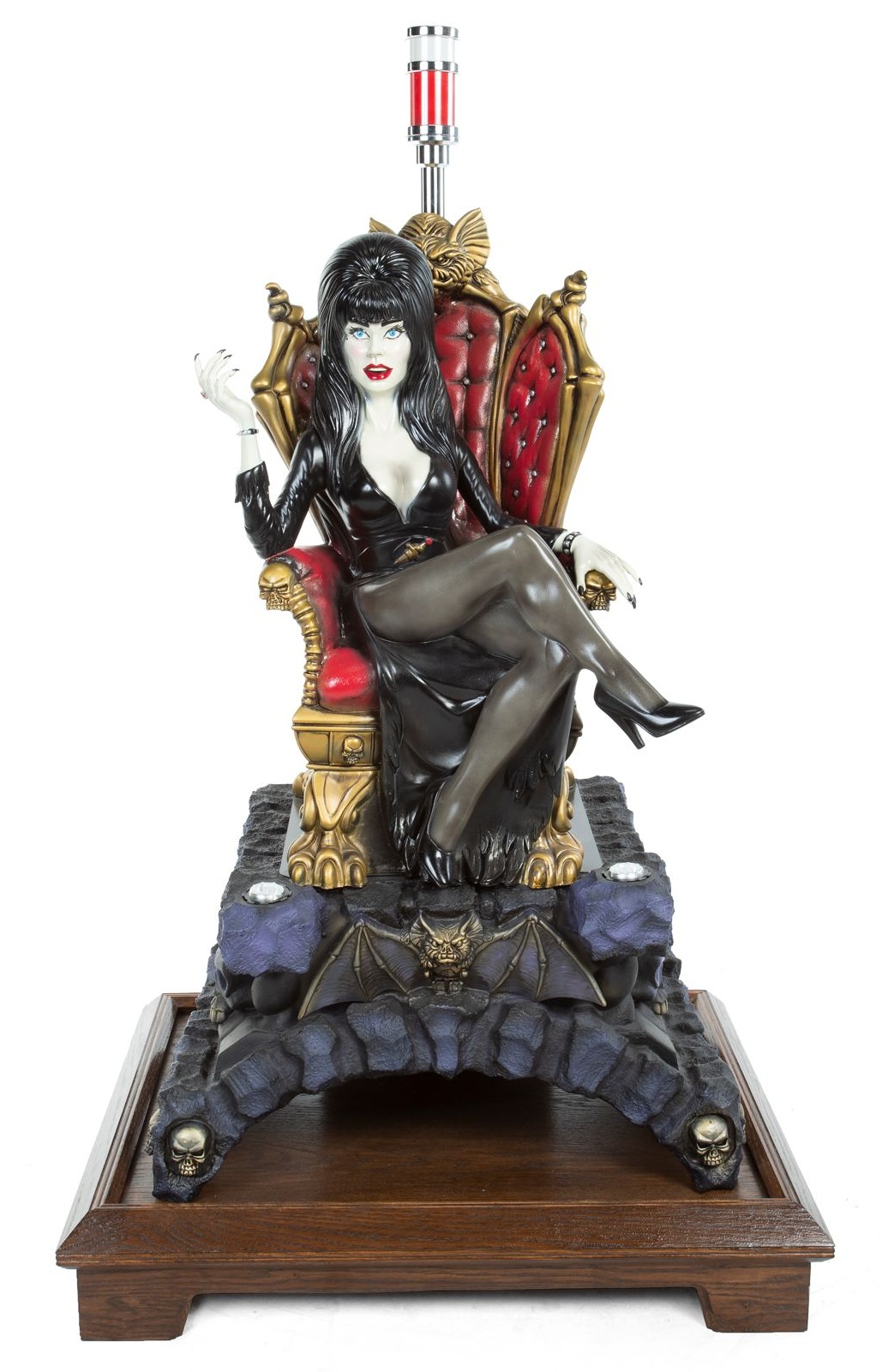 A figural light-up slot machine topper made of molded plastic in the form of Elvira sitting atop a red chair on a base decorated with bats and skulls, further atop a wooden stand. Light now not working as piece is not attached to a slot machine.