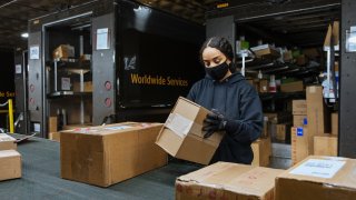 A Black woman stands in a warehouse, surrounded by packages in cardboard boxes. She wears a black hoodie, black gloves and a black COVID mask. A UPS truck is visible behind her, with the back doors to the truck's storage area open.