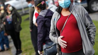 a pregnant woman wearing a face mask and gloves holds her belly as she waits in line