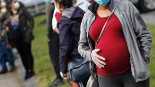 a pregnant woman wearing a face mask and gloves holds her belly as she waits in line