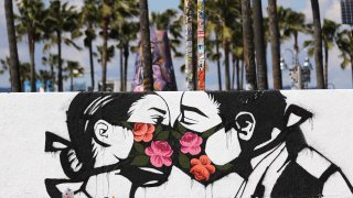VENICE, CALIFORNIA – MARCH 21: Palm trees stand behind a street art piece by artist Pony Wave depicting two people kissing while wearing face masks on Venice Beach on March 21, 2020 in Venice, California. California Governor Gavin Newsom issued a ‘stay at home’ order for California’s 40 million residents in order to slow the spread of coronavirus (COVID-19). Californians may still go to the beach without violating Newsom’s order as long as they maintain social distancing and adhere to other public health measures related to the coronavirus. (Photo by Mario Tama/Getty Images)