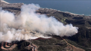 Aerial view of smoke and wildfires threatening an affluent oceanside neighborhood.