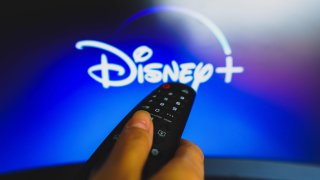 In this photo illustration, a hand holding a TV remote control in front of the Disney Plus logo on a TV screen.