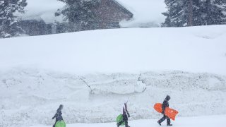 People walk past a snowbank piled up in front of condominiums as snow falls in the Sierra Nevada mountains from yet another storm system which is bringing heavy snow to higher elevations while further raising the snowpack on March 28, 2023 in Mammoth Lakes, California.