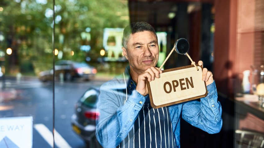 Mature man in his 40s holding open sign on door of business, looking through window, optimism, aspiration, resilience