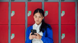 A medium close-up front view of a teenage girl who is sending text messages to a friend and checking her social media as she waits in the corridor of the high school she attends. 