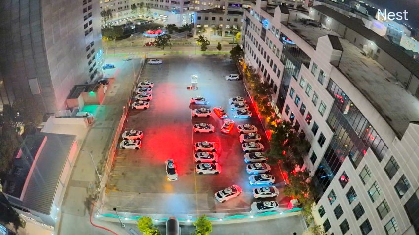 A video from a surveillance camera in a building shows a parking lot below at night time with several vehicles with lights on backing into spots