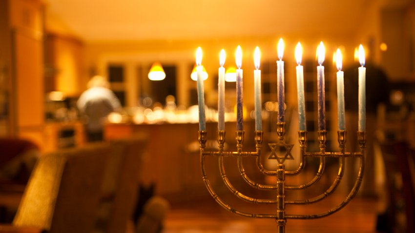 Lit candles in a menorah with a women working in the kitchen in the background, commonly used during the Jewish celebration of Hanukkah.