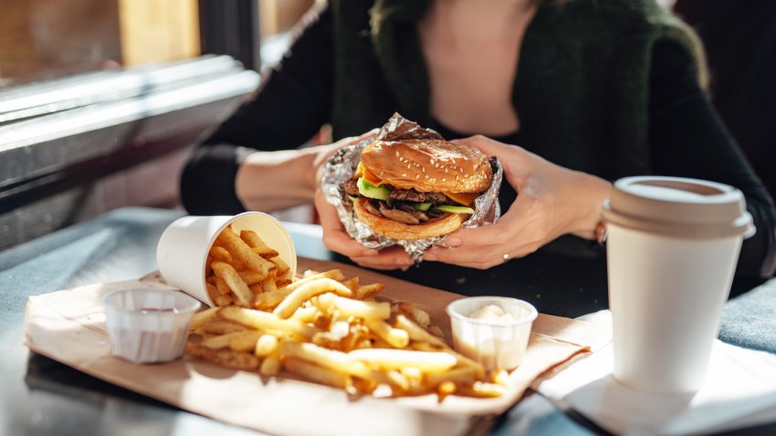 Woman holding large burger in front of a plate of fries and large drink.