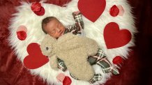 A newborn sleeps on a white heart-shaped pillow covered with smaller red hearts.