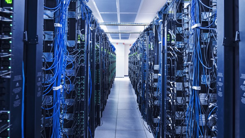 This image shows a long, well-organized corridor inside a modern data center. Rows of server racks line both sides of the corridor, their front panels lit with bright blue indicator lights. The reflective, smooth floor mirrors the overhead linear light fixtures, creating a symmetrical, high-tech visual of an IT infrastructure facility. The space is clean, empty, and designed to house critical digital storage and network hardware.
